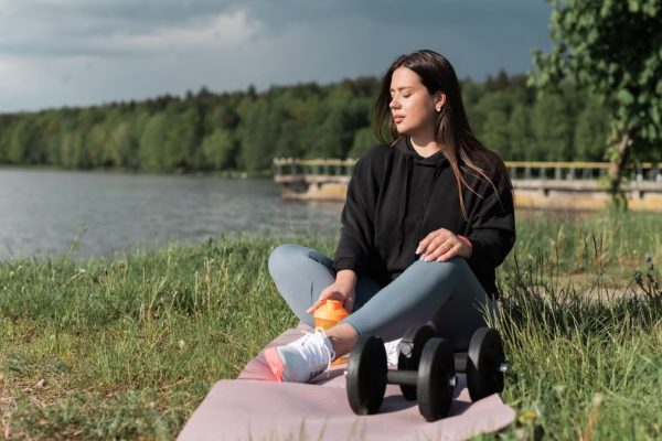 Woman sitting on yoga mat beside a lake with exercise equipment, enjoying a calm outdoor workout.
