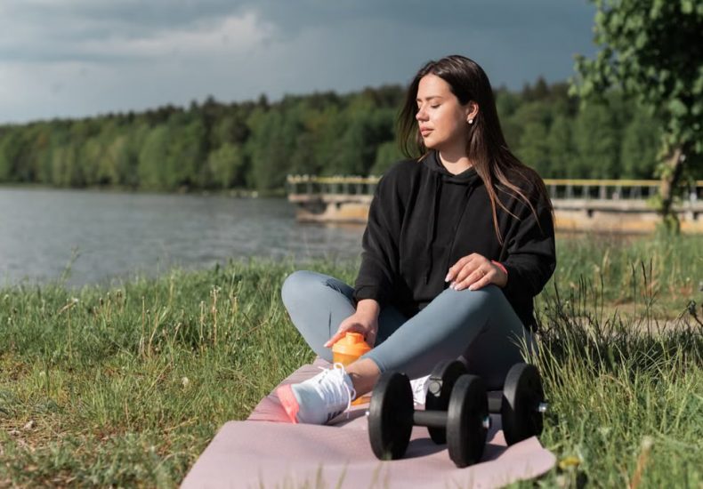 Woman sitting on yoga mat beside a lake with exercise equipment, enjoying a calm outdoor workout.