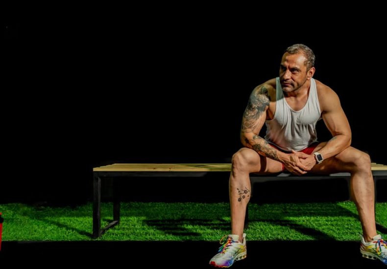 A muscular, tattooed man seated on a bench in a gym, depicting modern fitness lifestyle.